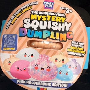 May include: A round cardboard packaging for a Mystery Squishy Dumpling toy. The front features text including "The Original Viral Mystery Squishy Dumpling," "Ages 3+," and "Pink Holographic Edition!" with illustrations of colorful, smiling dumplings.