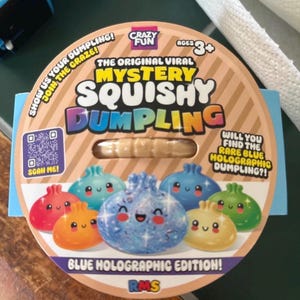 May include: A round cardboard packaging for a Mystery Squishy Dumpling toy. The label features the text "The Original Viral Mystery Squishy Dumpling" and "Blue Holographic Edition!" with colourful illustrations of various small, smiling dumpling characters.