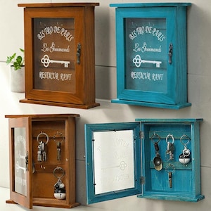 May include: Four rectangular wooden wall-mounted key cabinets with glass doors. Two are brown and two are teal. Each door features white text reading BISTRO DE PARIS Les Gouimands RESTAURANT and a key graphic. The cabinets contain metal hooks for keys.