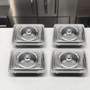 May include: Four square stainless steel food warmers with glass lids and silver knobs. Each unit features a polished metal base with an embossed decorative border pattern around the central circular glass insert, providing a clear view inside.