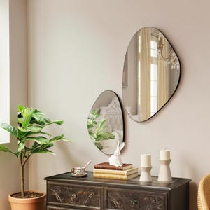 Può includere: Due specchi da parete con forme organiche irregolari e sottili cornici nere. Gli specchi riflettono l'interno di una stanza, inclusi una testiera bianca e un lampadario. Sono posizionati sopra una cassettiera in legno scuro.