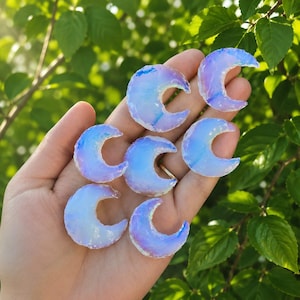 Pode incluir: Seis pedras em forma de meia-lua com um acabamento translúcido e iridescente. As pedras exibem tons variáveis de azul pálido, violeta suave e branco leitoso, refletindo a luz com uma qualidade brilhante e opalescente nas suas superfícies.