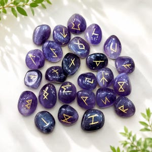 Peut inclure: Une collection de pierres d'améthyste polies d'un violet profond, chacune gravée d'un symbole runique doré unique. Les pierres lisses et arrondies sont disposées en grappe, mettant en valeur les variations naturelles des teintes violettes.