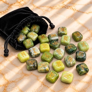 May include: A black velvet drawstring pouch containing and surrounded by a collection of small, smooth, square-shaped green stones. Each stone features a unique gold-coloured engraved rune symbol on its top surface, varying in shades of light to dark green.