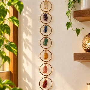 May include: A vertical wall hanging featuring seven gold-toned metal rings linked together in a chain. Each ring contains a single raw, wire-wrapped gemstone in a different colour, including clear, purple, blue, teal, yellow, pink, and red.