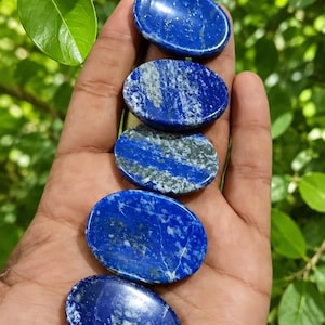 Puede incluir: Cinco piedras preciosas de lapislázuli de forma ovalada con un acabado pulido. Cada piedra muestra un color azul intenso con inclusiones naturales de calcita blanca y motas de pirita dorada distribuidas por las superficies lisas.