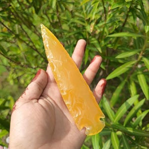 May include: A translucent yellow stone arrowhead with a sharp, pointed tip and a flaked, textured surface. The object features a notched base and a smooth, glossy finish, displaying a vibrant golden hue that catches the natural light.