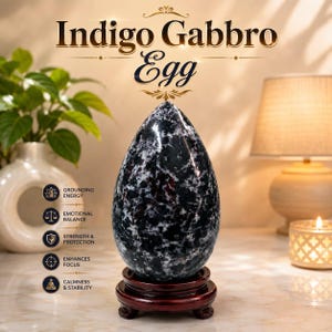 May include: An egg-shaped Indigo Gabbro stone with a polished, mottled black and white surface, resting on a circular dark wood stand. The text "Indigo Gabbro Egg" appears above, with a list of five attributes including grounding energy.