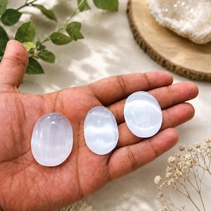 Könnte beinhalten: Drei glatte, oval geformte Selenit-Handschmeichler mit einem durchscheinenden, milchig-weißen Aussehen und einem reflektierenden, perlmuttartigen Glanz. Die Steine variieren leicht in der Größe und zeigen eine faserige, schimmernde Textur.