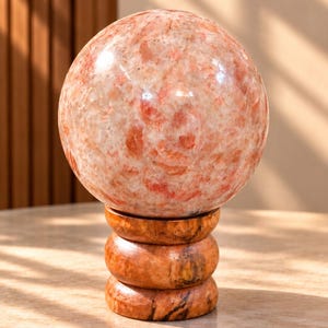 Peut inclure: Une sphère en pierre de soleil polie avec une texture cristalline tachetée de pêche, d'orange et de crème. La sphère repose sur un support en bois cylindrique présentant un grain brun naturel et un design à anneaux empilés arrondis.