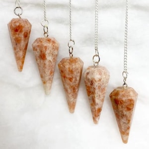 Può includere: Cinque pendoli in pietra del sole, ognuno caratterizzato da una pietra sfaccettata a forma di cono con motivi maculati arancioni e bianchi. Ogni pietra è sospesa a una catenella in metallo argentato fissata a un piccolo anello circolare.
