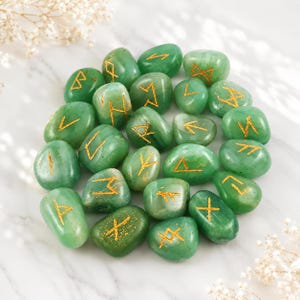 Op de afbeelding: Een verzameling gladde, gepolijste groene stenen, elk gegraveerd met een uniek gouden runensymbool. De stenen zijn afgerond en variëren enigszins in vorm en grootte, met een levendige groene tint en natuurlijke variaties in kleur en textuur.