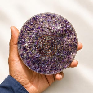 Könnte beinhalten: Ein kreisförmiger Untersetzer oder Briefbeschwerer aus Kunstharz, der mit zahlreichen kleinen, violetten Amethyst-Kristallsplittern eingebettet ist. Ein dünner, dunkler Kupferdraht ist spiralförmig gewickelt und in der Mitte des transparenten, glatten Objekts platziert.