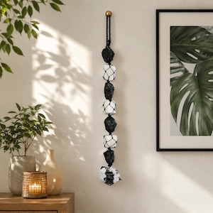 Puede incluir: Un colgante de pared vertical con un cordón negro trenzado que sostiene piedras negras rugosas y piedras blancas lisas alternadas dentro de una red negra, terminado con un grupo de cristales pequeños transparentes y negros.