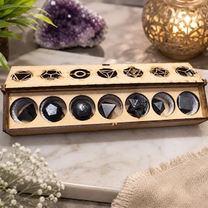 Puede incluir: Una caja rectangular de madera con tapa abatible que contiene siete formas geométricas poliédricas negras. La tapa presenta diseños cortados con láser que corresponden a cada forma, incluyendo una esfera, un cubo y varias pirámides.