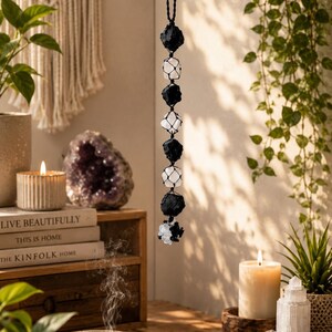 Può includere: Una decorazione da parete verticale con cristalli alternati di tormalina nera grezza e quarzo trasparente, racchiusi in una rete di corda macramè nera. L'oggetto è sospeso a un cordino nero e termina con un grappolo di pietre più piccole.