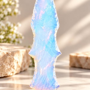 Könnte beinhalten: Eine durchscheinende, irisierende blaue Speerspitze mit einer scharfen Spitze und gezackten Kanten. Die Oberfläche des Objekts zeigt eine schimmernde, opalisierende Qualität, wobei sich das Licht in sanften Rosa- und Violetttönen auf der Form spiegelt.