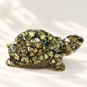 Op de afbeelding: Een schildpadfiguur gemaakt van talloze kleine, hoekige stukjes metaalachtig goud en donkergrijze steen. Het schild en het lichaam hebben een mozaïekachtige textuur met reflecterende oppervlakken, wat zorgt voor een gefacetteerd uiterlijk.