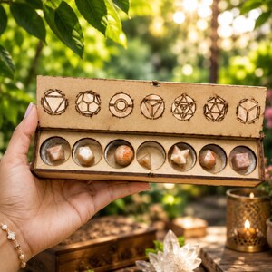 Puede incluir: Una caja rectangular de madera con tapa abatible que contiene siete dados poliédricos geométricos distintos hechos de piedra marrón claro. La tapa presenta siete iconos circulares grabados con láser que representan las formas.