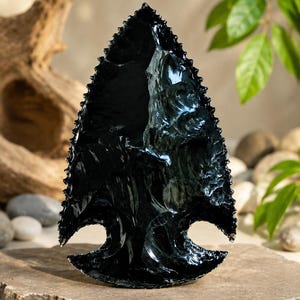 Könnte beinhalten: Eine schwarze Obsidian-Pfeilspitze mit einer scharfen Spitze und gezackten Kanten. Die Oberfläche des Steins ist glatt und reflektierend und weist eine glänzende, glasartige Textur mit einer ausgeprägten dreieckigen Form auf.
