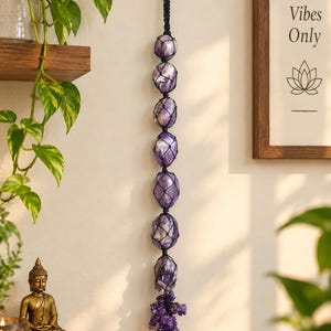 May include: A vertical wall hanging featuring seven polished purple amethyst stones encased in a black macrame cord net. The stones are arranged in a single column, ending with a cluster of smaller, raw amethyst crystals at the bottom.