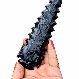Könnte beinhalten: Eine lange, spitze Obsidianklinge mit glänzend schwarzer Oberfläche und gezackten Kanten. Der Stein hat eine scharfe, zulaufende Spitze und eine breite, eingekerbte Basis, die eine glatte, reflektierende Oberfläche mit muscheligen Bruchmustern zeigt.