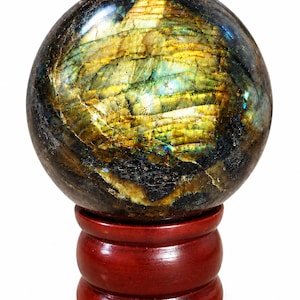 May include: A polished labradorite sphere displaying iridescent flashes of gold, yellow, and blue across its dark, fractured surface. The stone rests securely on a smooth, circular, dark reddish-brown wooden stand against a plain white background.