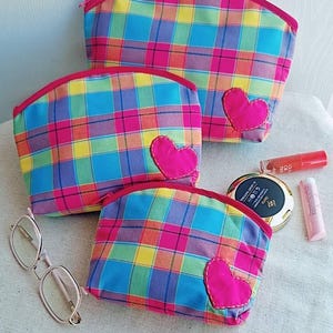 May include: Three zippered pouches in varying sizes featuring a vibrant plaid pattern of pink, blue, yellow, and green. Each pouch is accented with a single, stitched-on bright pink fabric heart. Also included are eyeglasses, a compact, and lip products.