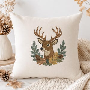 May include: A square off-white throw pillow featuring a central embroidered design of a brown deer head with antlers, framed by a wreath of green pine needles, brown pinecones, and tan oak leaves, centered on the front of the pillow cover.