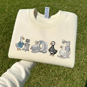 Peut inclure: Un sweat-shirt à col rond couleur crème avec une broderie représentant quatre canards dans un environnement médical. Le motif inclut un canard avec un stéthoscope, un canard près d'un scanner CT, un canard en costume de squelette et un canard près d'une machine à rayons X.