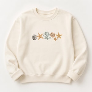 Op de afbeelding: Een crèmekleurige sweater met ronde hals en een centrale grafische print van diverse zeedieren, waaronder twee oranje zeesterren, een lichtblauwe koraaltak en twee bruine schelpen die horizontaal op de borst zijn gerangschikt.