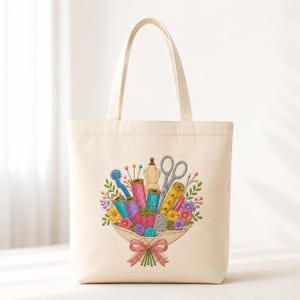 May include: A cream-colored canvas tote bag featuring a colorful embroidered bouquet made of sewing supplies. The design includes spools of thread, scissors, a measuring tape, a thimble, buttons, and pins, all tied together with a pink ribbon.