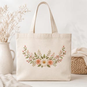 May include: A cream-colored canvas tote bag featuring a central embroidered floral arrangement. The design displays a symmetrical garland of green leaves, small pink buds, and a cluster of white and peach-colored daisy-like flowers.