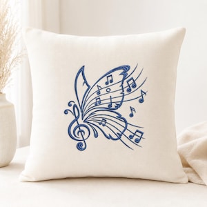 Könnte beinhalten: Ein quadratisches cremefarbenes Zierkissen mit einer mittigen, dunkelblauen Stickerei in Form eines Schmetterlings. Der Schmetterlingsflügel besteht aus Notenlinien, einem Violinschlüssel und verschiedenen Noten, die über den Flügel verteilt sind.