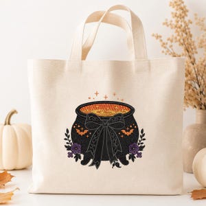 May include: A natural-colored canvas tote bag featuring an embroidered black cauldron with an orange swirling liquid interior, a large black bow, small orange bats, and purple flowers. The design includes small orange star accents above the cauldron.