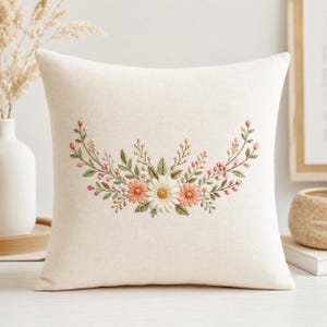 May include: A square off-white throw pillow featuring a central embroidered floral arrangement. The design includes a large white daisy, two smaller peach-colored flowers, and delicate green leafy stems with tiny pink buds extending outward.