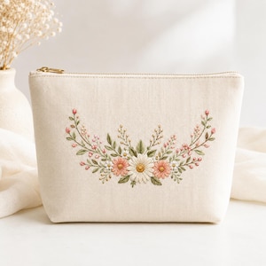 May include: A cream-colored canvas pouch featuring a central floral embroidery design. The embroidery depicts a symmetrical arrangement of white and peach-colored daisies, green leaves, and small pink buds, secured with a gold-toned metal zipper.