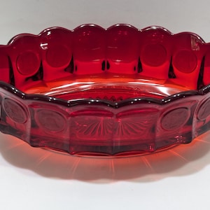 Puede incluir: Un cuenco de vidrio rojo rubí con borde festoneado y un patrón decorativo. El cuenco tiene una serie de diseños circulares y en forma de abanico alrededor de los lados. El cuenco tiene forma ovalada y se apoya sobre una superficie blanca.