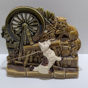 Puede incluir: Una figura de cerámica que representa un gato y un perro con una rueca. El perro es blanco con manchas marrones y está con correa. El gato es marrón. La figura es principalmente marrón y verde.