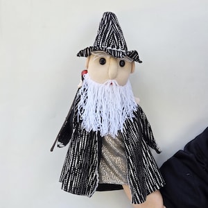Puede incluir: Una muñeca de mago hecha a mano con un sombrero y abrigo a rayas blancas y negras. La muñeca tiene una larga barba blanca, una túnica dorada y un acento rojo. La muñeca está sujeta por una persona.
