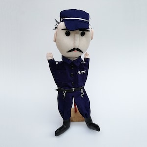 Puede incluir: Un títere de mano de fieltro azul vestido de policía. El títere tiene una cara blanca con un bigote negro y un sombrero azul con una banda plateada. El títere lleva un uniforme azul con un cinturón negro y llaves plateadas. El uniforme tiene la palabra "POLICE" impresa en el pecho.