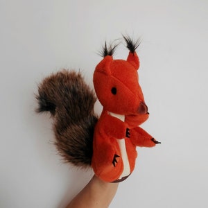 May include: Orange and brown hand puppet of a squirrel with a fluffy tail. The puppet has a white face with black eyes and a black nose.