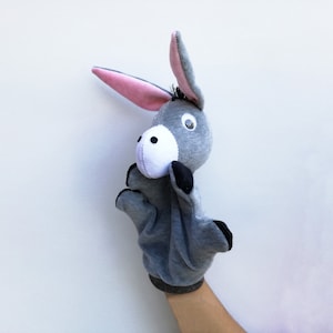 May include: A gray donkey puppet with pink ears and black eyes. The puppet is being held up by a hand.