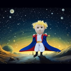 Peut inclure: Une poupée en peluche d'un personnage du "Petit Prince" avec des cheveux jaunes, une cape rouge et bleue et un pantalon blanc. La poupée est placée devant un fond de ciel étoilé avec un paysage.