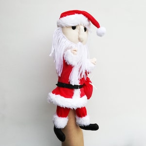 Peut inclure: Une marionnette de Père Noël en peluche rouge et blanche avec une barbe blanche et une ceinture noire. La marionnette porte un chapeau rouge avec un pompon blanc. La marionnette tient un sac rouge dans sa main droite.