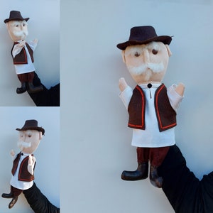 Puede incluir: Títere hecho a mano de un anciano con sombrero marrón, chaleco con ribete naranja, camisa blanca y pantalón oscuro. El títere tiene bigote blanco y posa con los brazos en alto. El títere está sujeto por una mano.