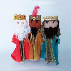 Puede incluir: Tres marionetas de mano vestidas como los tres reyes magos. Las marionetas están hechas de fieltro y tienen caras bordadas. Los reyes magos llevan túnicas y coronas tradicionales. La marioneta de la izquierda lleva una túnica roja, la marioneta del centro lleva una túnica dorada y la marioneta de la derecha lleva una túnica azul.