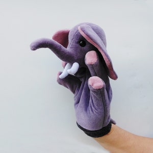 May include: Purple plush elephant hand puppet with pink ears and white tusks. The puppet is worn on a hand and has a black wrist band.