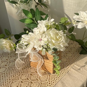 Arreglo floral artificial hecho a mano con flores blancas en maceta de madera, ideal para decorar la mesa de una boda o despedida de soltera.