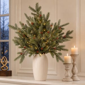 May include: A white ribbed ceramic vase holds a lush arrangement of artificial evergreen branches featuring small red berries and integrated warm white fairy lights. Two cream-colored pillar candles sit in weathered wooden candle holders nearby.